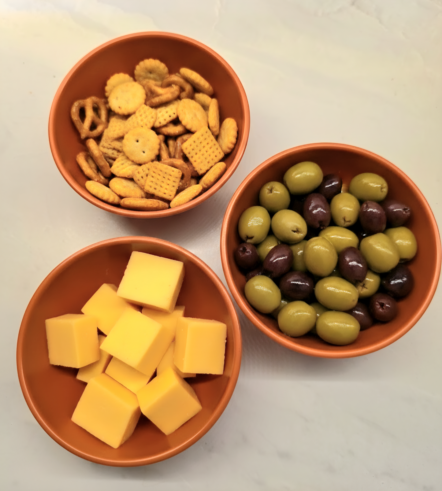 Three terracotta bowls of snacks including crackers, cheese cubes, and olives on a light surface.