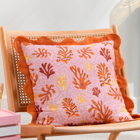 Mediterranean-style coral orange accent cushion styled on a wooden wicker chair for a coastal outdoor aesthetic.