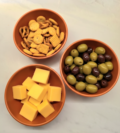 Three terracotta bowls of snacks including crackers, cheese cubes, and olives on a light surface.